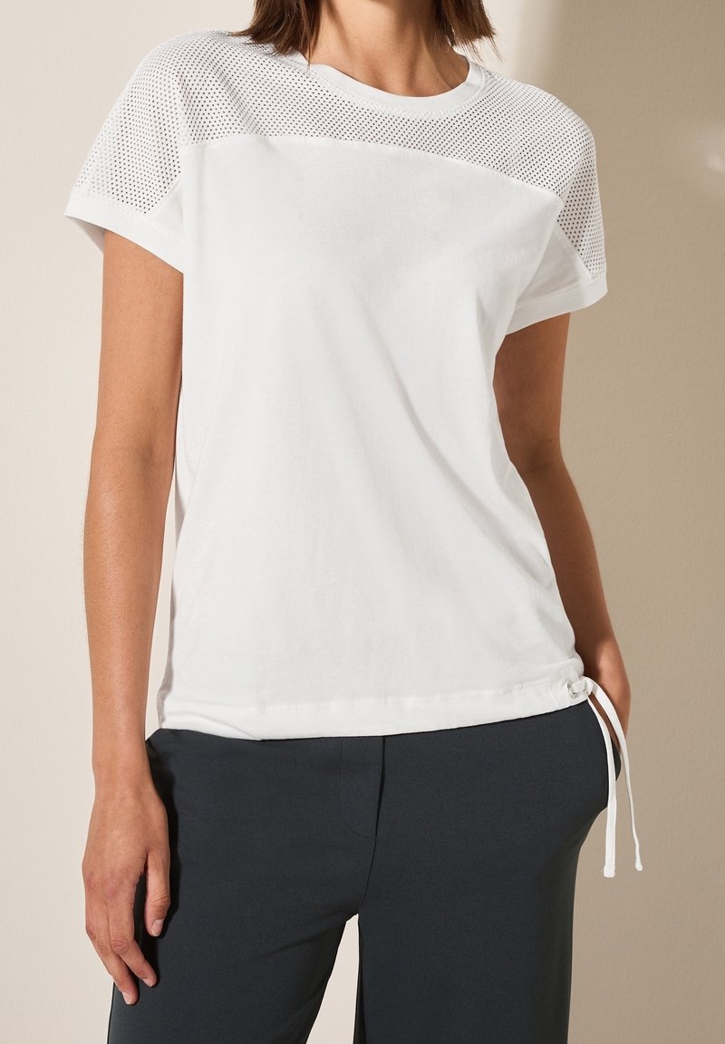 Femme portant un haut blanc à manches courtes avec épaules perforées et ourlet à cordon, associé à un pantalon noir sur fond beige.