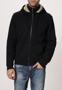 Uomo che indossa una felpa blu navy con zip e interno della cappuccio in pile beige, jeans blu sbiaditi, con una mano in tasca.