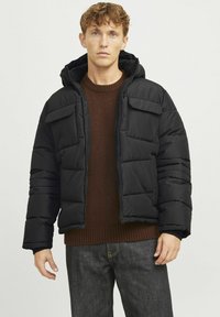 Pufferjakke i sort med lynlås, to brystlommer og hætte. Båret over en brun sweater med ribbet tekstur.
