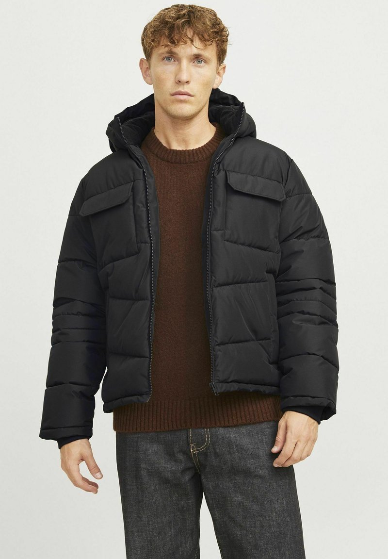 Pufferjakke i sort med lynlås, to brystlommer og hætte. Båret over en brun sweater med ribbet tekstur.