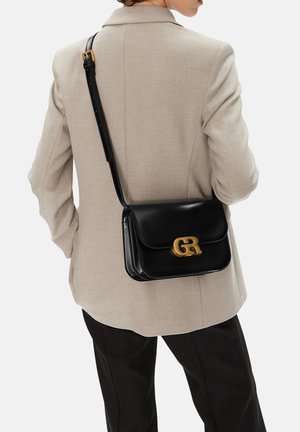 Osoba ubrana w beżową marynarkę i czarne spodnie z czarną skórzaną torebką na ramię z zapięciem z złotym logo GR.