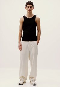 Schwarzes ärmelloses Tanktop, lockere cremefarbene Hose und weiße Sneaker. Das Model steht vor einem schlichten hellen Hintergrund.