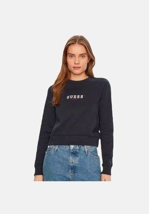 Felpa blu navy con scollatura rotonda e polsini a costine, con il logo "GUESS?" in colori contrastanti sul davanti. Abbinata a jeans di denim.
