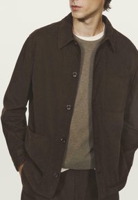 Donkerbruin jasje met een knoopsluiting, grote opgestikte zakken en een klassieke kraag, gecombineerd met een grijze trui eronder.