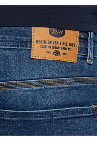 Jeans de mezclilla en azul oscuro con un acabado texturizado. Presentan una etiqueta de cuero marrón claro que dice "Denim Driven Since 1989" y costuras de color amarillo en contraste.