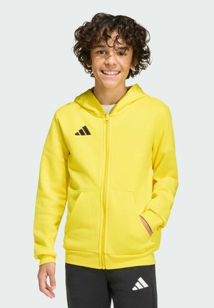 Lächelnde junge Person trägt einen leuchtend gelben Kapuzenpullover mit Reißverschluss und schwarze Hosen mit Adidas-Logos, steht mit einer Hand in der Tasche.