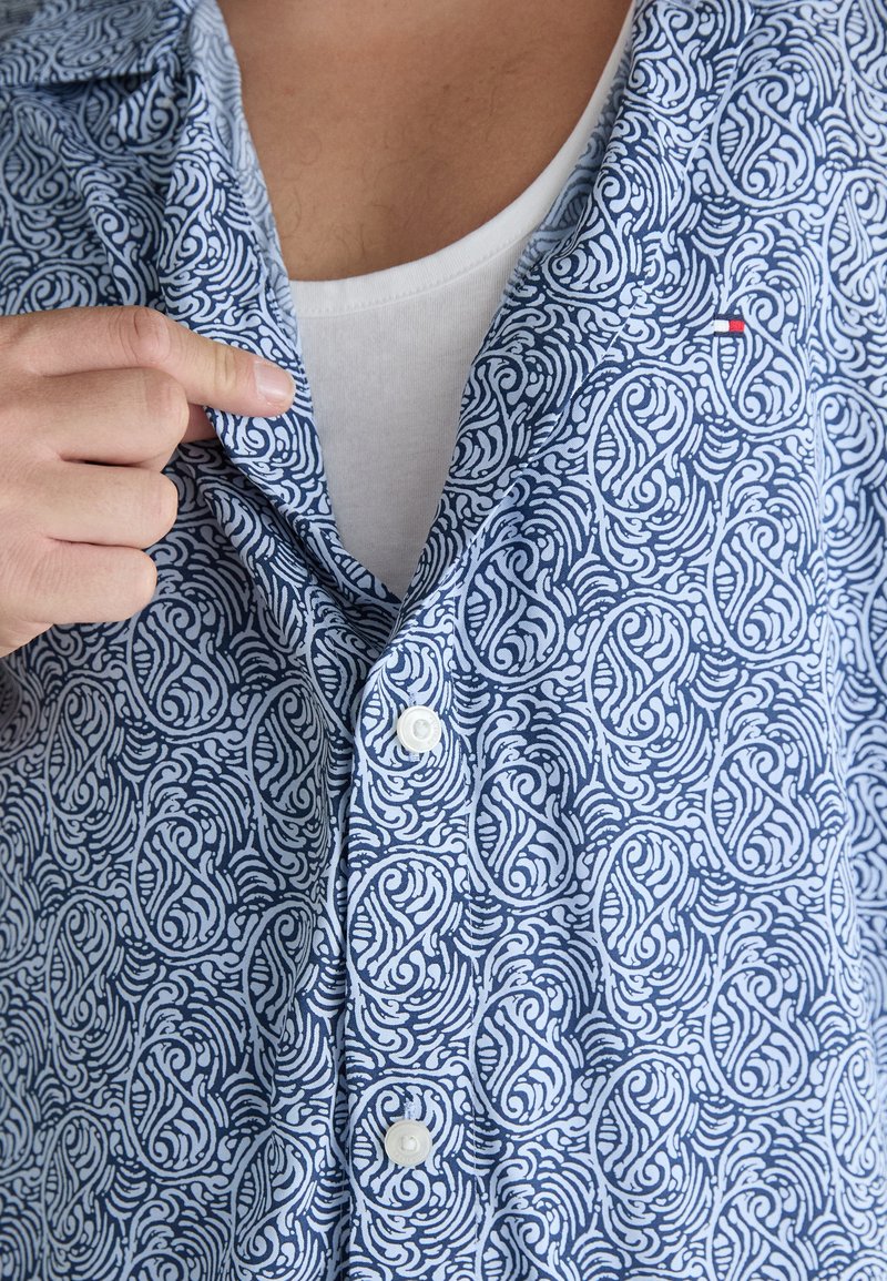 Chemise à manches courtes, boutonnée, en tissu bleu avec un motif complexe de tourbillon blanc. Un bouton blanc visible près du col.
