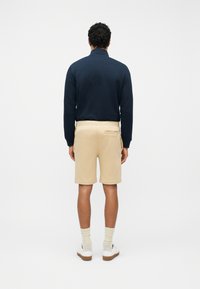 Άνδρας που φοράει μπλε μακρυμάνικη μπλούζα, μπεζ σορτς με πίσω τσέπη, κρεμ κάλτσες και λευκά αθλητικά παπούτσια, στέκεται με την πλάτη στο φακό μπροστά σε απλό φόντο.