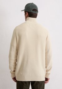 Roomkleurige geribbelde gebreide trui met een hoge kraag, gecombineerd met een groene corduroy pet. Het ontwerp heeft lange mouwen en een losse pasvorm.