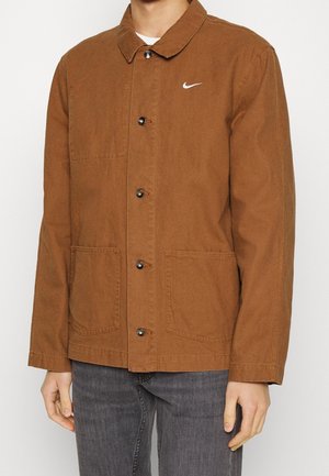 Bruine katoenen jas met een puntkraag, knoopsluiting, twee voorzakken en een wit Nike-logo op de linker borst. Semi-gestructureerd ontwerp.