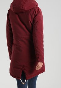 Parka acolchada burdeos con capucha, que presenta una cintura ceñida, cordón ajustable en el dobladillo y un detalle triangular en la parte trasera.