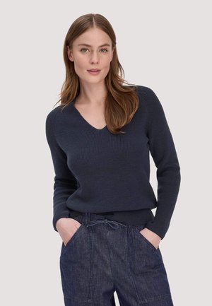 Junge Frau mit hellbraunen Haaren trägt einen marineblauen V-Ausschnitt-Strickpullover und blaue Jeanshose, steht mit den Händen in den Taschen vor einem schlichten Hintergrund.