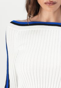 Hvit ribbestrikket genser med av-shoulder design og aksentstriper i blått og svart ved halsen. Myk tekstur og tettsittende fasong.