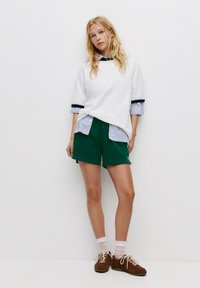 Weißer übergroßer Pullover mit marineblauen Akzenten, über einem hellblauen gestreiften Hemd getragen, kombiniert mit grünen Shorts und braunen Turnschuhen.