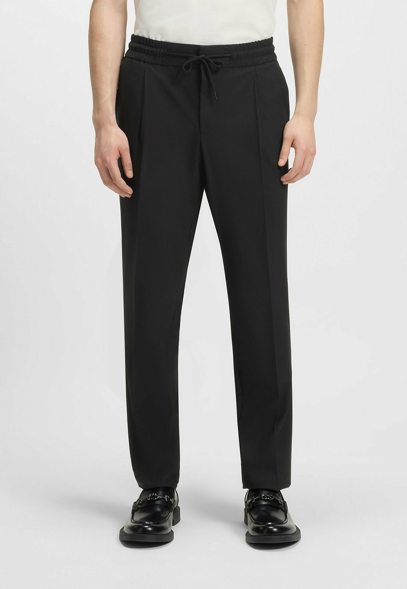 Black tailored pants with an elastic waistband and drawstring. Features two side pockets and a smooth, sleek fabric finish.
