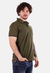 Camisa de polo verde oliva con mangas cortas, cuello y puños negros, tapeta de tres botones y detalle de logo bordado en el pecho.