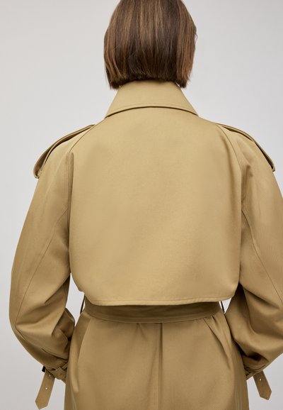 Cappotto trench beige con un design corto, ampio colletto e spalline. Tessuto liscio con vita accentuata da una cintura e maniche lunghe.