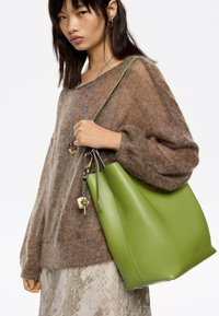 Femme portant un pull en maille marron et une jupe à motifs, tenant un grand sac fourre-tout en cuir vert avec un accessoire porte-clés.