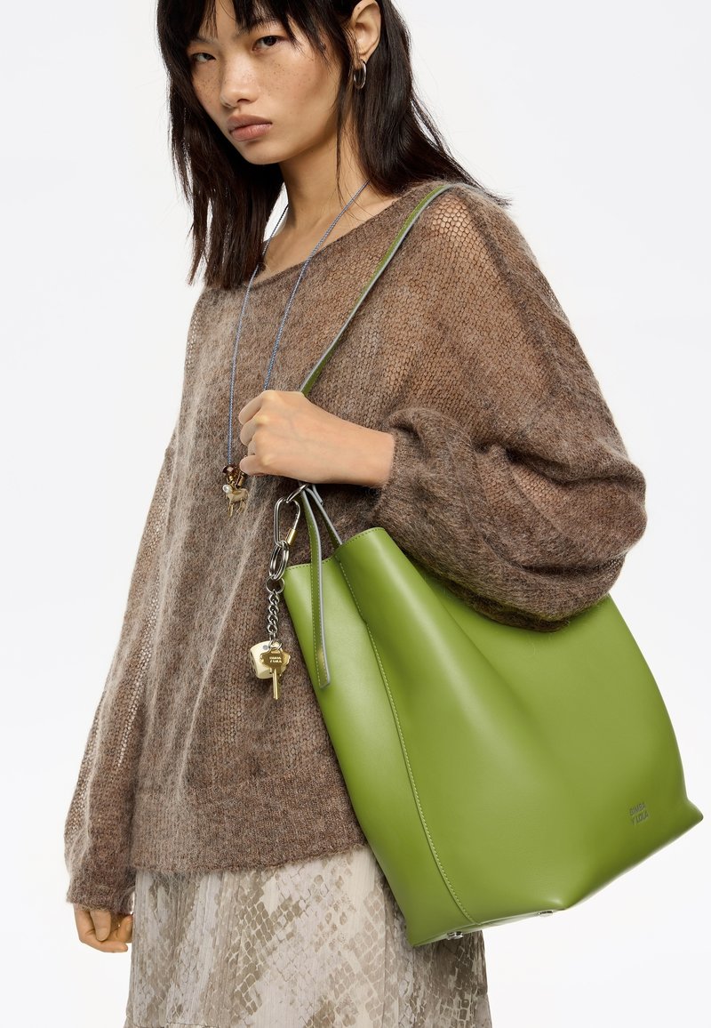 Femme portant un pull en maille marron et une jupe à motifs, tenant un grand sac fourre-tout en cuir vert avec un accessoire porte-clés.