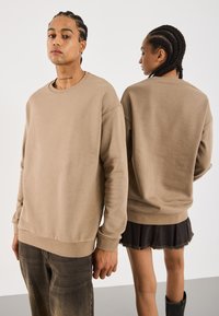 To personer med flettet hår har på seg beige gensere; den ene står vendt mot kameraet med jeans, mens den andre viser ryggen iført et svart skjørt og støvler.