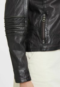 Giacca in pelle nera con dettagli trapuntati sulle spalle, tasca laterale con zip e texture liscia. Presenta un design aderente e un polsino arrotondato.