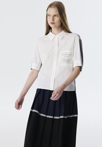 Camisa blanca de manga corta con cuello de botones y un bolsillo de parche. Combinada con una falda de tablas en azul marino y negro con un borde de encaje.