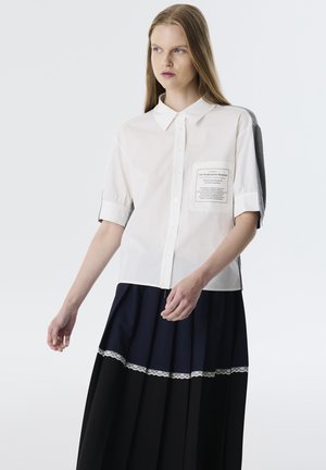 Camisa blanca de manga corta con cuello de botones y un bolsillo de parche. Combinada con una falda de tablas en azul marino y negro con un borde de encaje.
