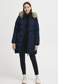 Mornarska pufasta jakna s odstranljivim umetnim krznom na kapuci, z značilnim prešitim dizajnom, sprednjim zadrgo in stranskimi žepi. Nosi se čez črn outfit.
