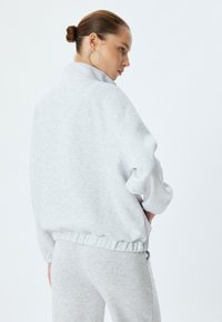 Sudadera de gran tamaño en gris claro con cuello alto y cinturilla elástica, combinada con pantalones grises a juego. Material suave y texturizado.
