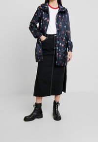 Giacca impermeabile blu navy con stampa floreale multicolore, polsini elasticizzati, abbinata a una T-shirt bianca e una gonna di denim nera, stivaletti neri.