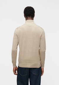 Άνδρας με πλάτη φοράει μπεζ πουλόβερ μακριάς μανίκας και σκούρο μπλε τζιν μπροστά σε απλό φωτεινό φόντο.