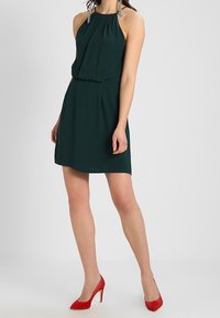 Robe dos nu vert foncé avec un corsage plissé et une taille cintrée. Portée avec des chaussures à talons rouges. Le tissu semble lisse et léger.