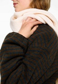 Beige texturerad scarf hängande runt en mörkbrun och blå zigzag-mönstrad kappa. Scarfen har ett mjukt, fluffigt utseende.