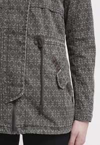Gray patterned jacket with a textured fabric, featuring a zipper, side pocket, and contrasting brown accents on the pockets and drawstring.