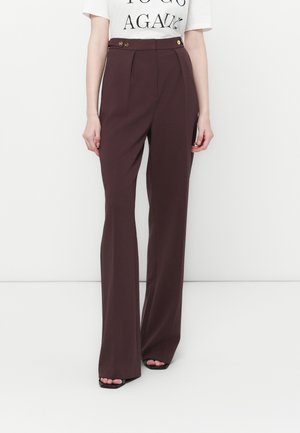 Woman wearing high-waisted, wide-leg dark plum trousers with gold button details and black open-toe heels, paired with a white printed top.