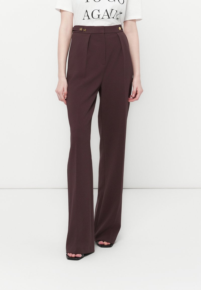 Femme portant un pantalon large taille haute de couleur prune foncé avec des détails de bouton doré et des talons ouverts noirs, associé à un haut blanc à motifs.