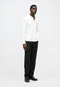 White long-sleeve polo shirt with a collar and small logo, paired with black trousers and black shoes, standing against a neutral background.