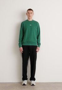 Sudadera verde con cuello redondo y pequeño logo blanco, combinada con pantalones negros y zapatillas deportivas grises, de pie contra una pared lisa.