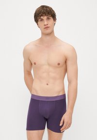 Boxers de algodão roxos com uma faixa suave na cintura, apresentando um logótipo discreto. O modelo posa sem camisa contra um fundo branco simples.