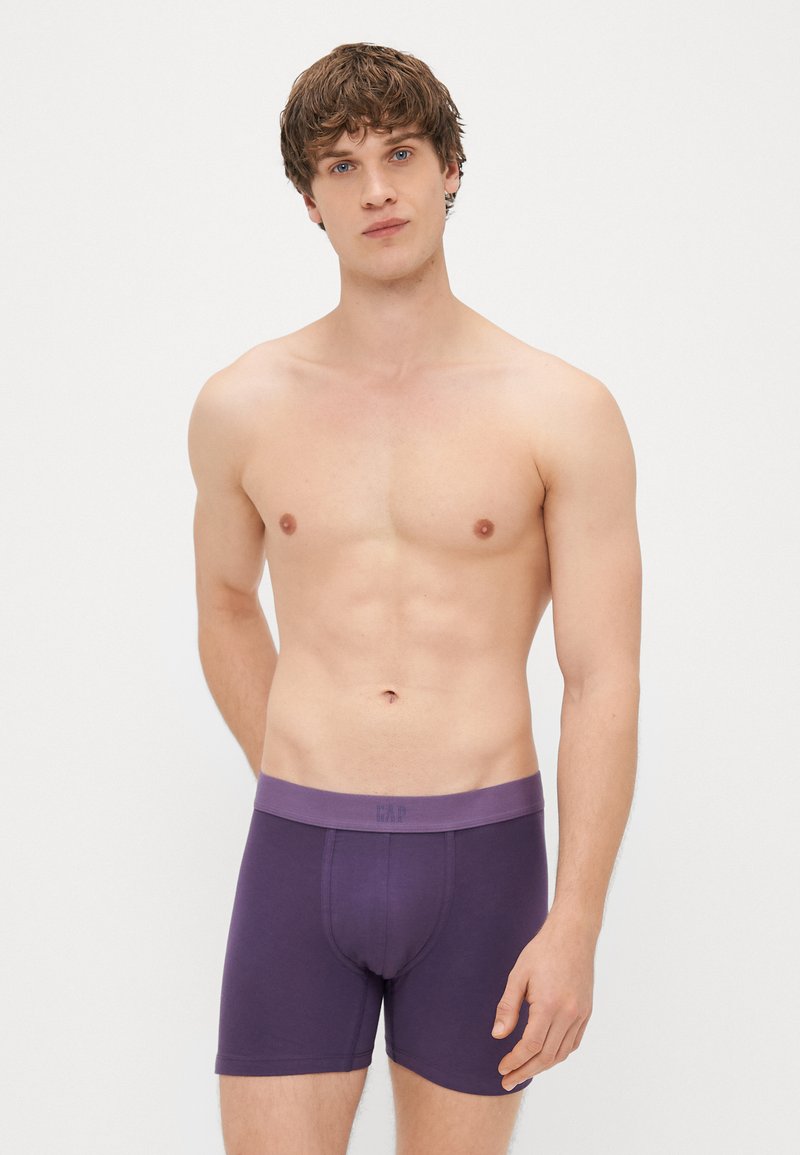 Boxers de algodão roxos com uma faixa suave na cintura, apresentando um logótipo discreto. O modelo posa sem camisa contra um fundo branco simples.