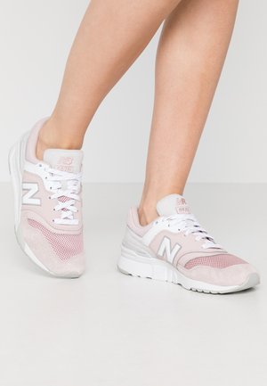 Osoba nosząca jasnoróżowe i białe sneakersy New Balance z siateczkowymi i zamszowymi detalami, stojąca na białej podłodze na tle jednolitego tła.