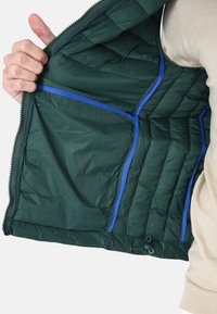 Interno della giacca a vento verde scuro con accenti blu, texture trapuntata, molteplici tasche e cordoncino all'orlo. Chiusura con zip.