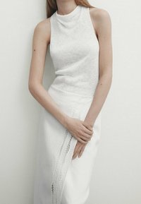 Biały top bez rękawów z wysokim kołnierzem, wykonany z miękkiego materiału, zestawiony z białą spódnicą z bocznym koronkowym detalem. Prosty i elegancki design.