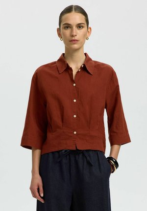 Naine, kes kannab roostevärvi nööpidega särki 3/4 varrukatega, tumesid nöörpükse, kuldseid rõngaskõrvarõngaid ja musta ning pruuni värvi käevõrusid.