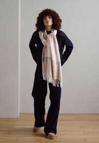 Tume meresinine kudumisel sweater, laiad mustad püksid ja hele roosa sall hallide ridade ja tuksuvate servadega. Puidust põrand, neutraalne seinakujundus.