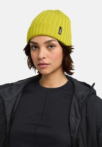 Gehaakte gele beanie met ribbelstructuur en een zwart label. Draag het met een zwarte, geperste jas over een zwarte T-shirt met ronde hals.