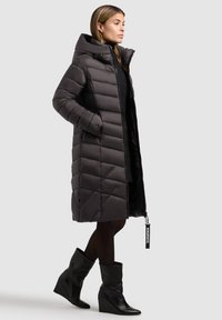Pufová bundá, tmavošedá, po kolená, diagonálne prešívaný vzor, vysoký golier, zips, bočné vrecká, oblečená s priliehavými čiernymi čižmami.
