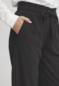 Juodos, aukšto liemens kelnės su lygaus paviršiaus, elastingu juosmeniu ir šonine kišene, su raišteliu priekyje.
