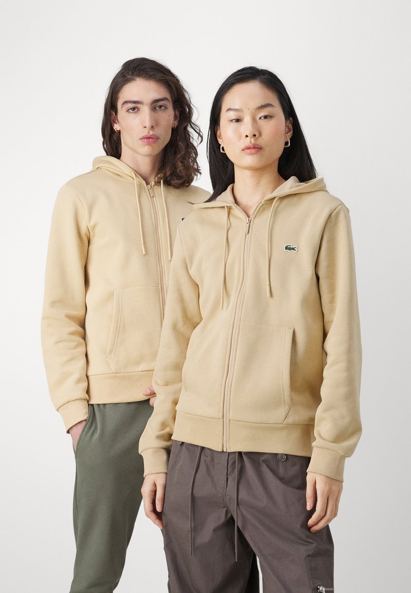 Twee modellen dragen beige ritshoodies met trekkoorden en voorzakken. De hoodies hebben het Lacoste-logo in het groen. Het mannelijke model combineert het met groene broeken, terwijl het vrouwelijke model cargo-stijl bruine broeken draagt.