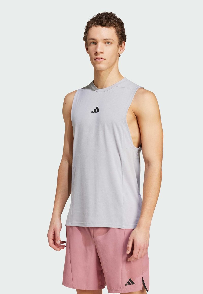 Ljusgrå ärmlös träningsöverdel med en Adidas-logotyp på bröstet. Har en avslappnad passform och en mjuk tygstruktur. Sammanförd med rosa shorts.